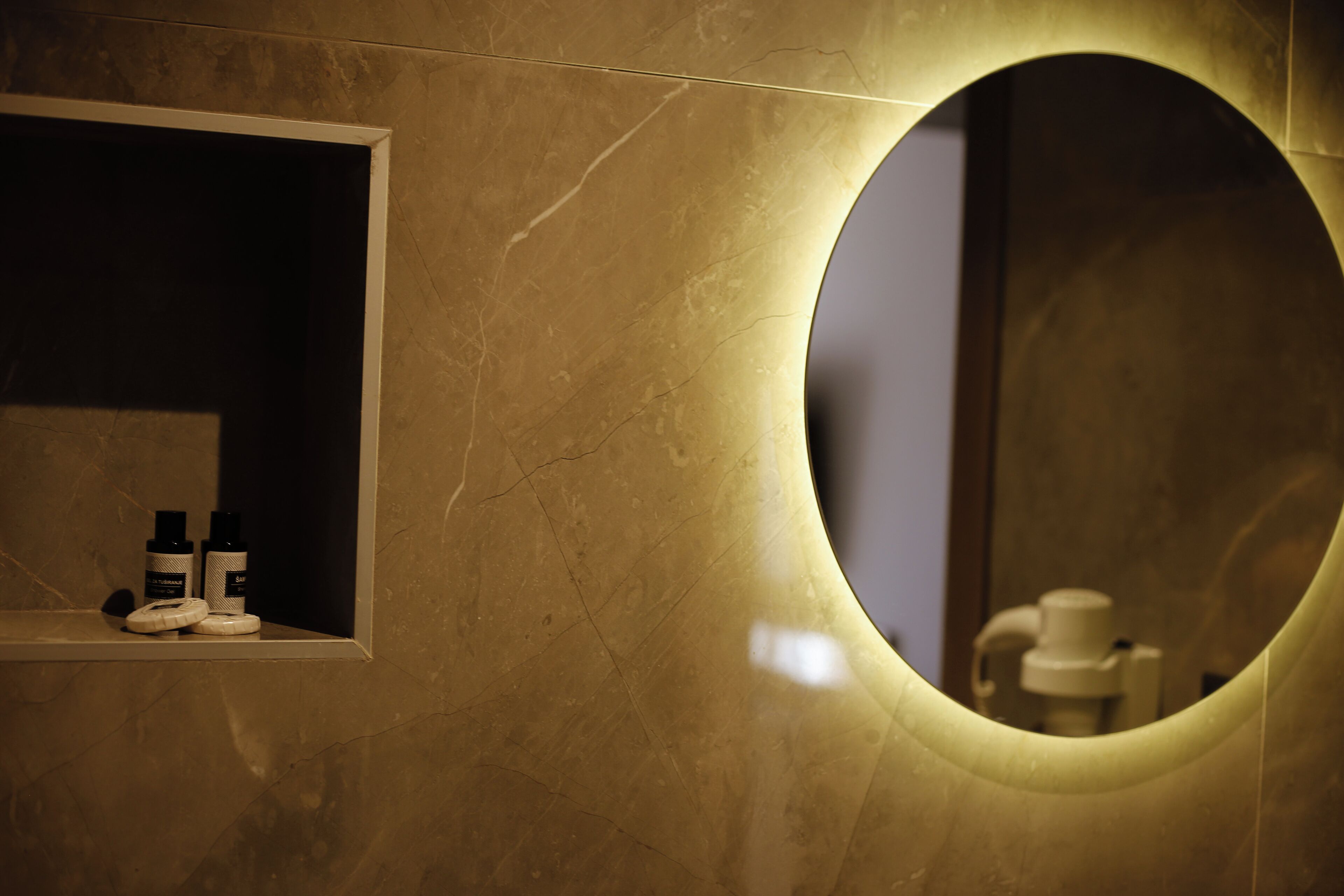 A bathroom with a marble wall, a round mirror, and a shelf with toiletries.