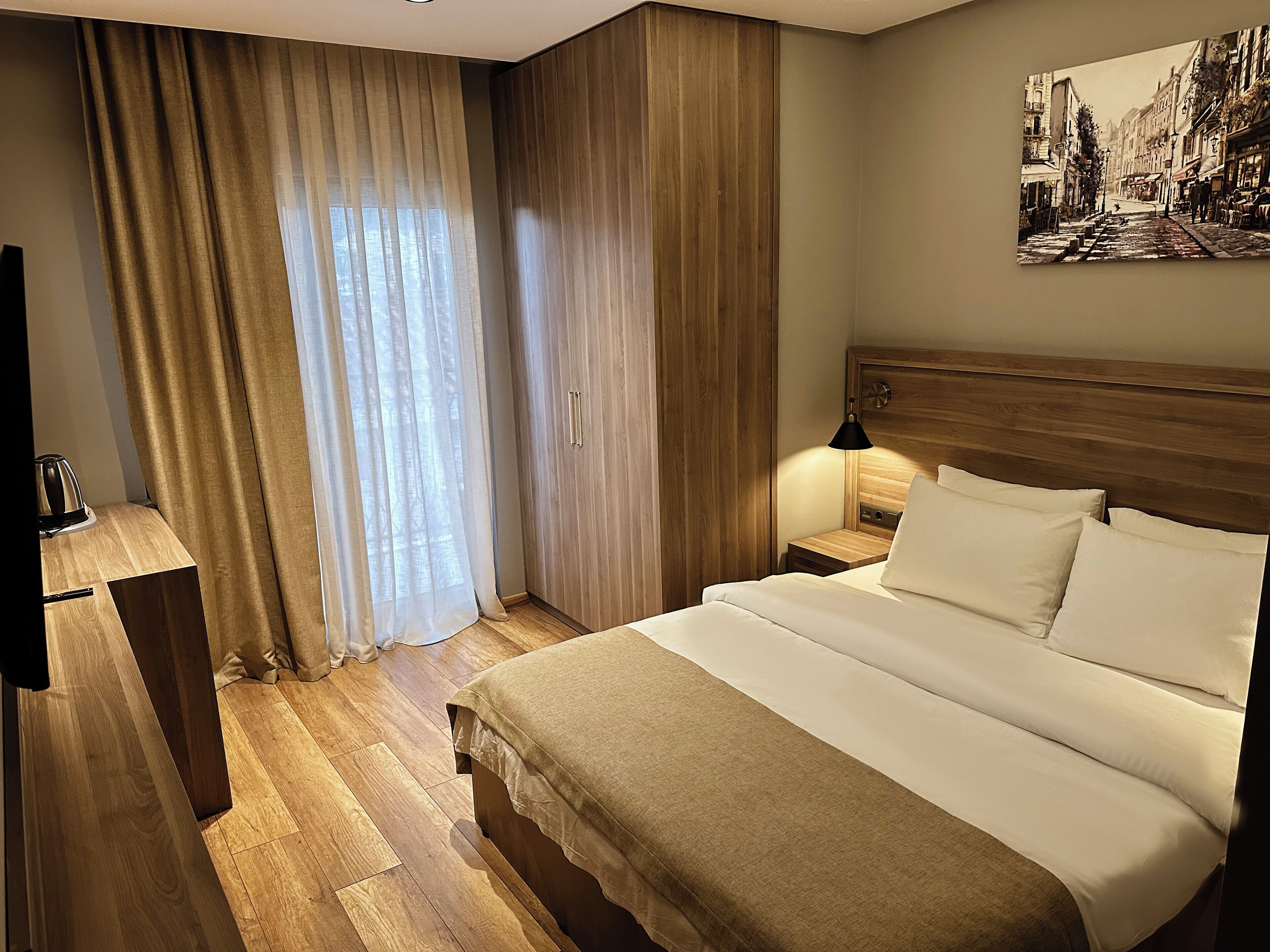 A hotel room with a wooden floor, a bed with white linens, a nightstand with a lamp, and a painting on the wall.