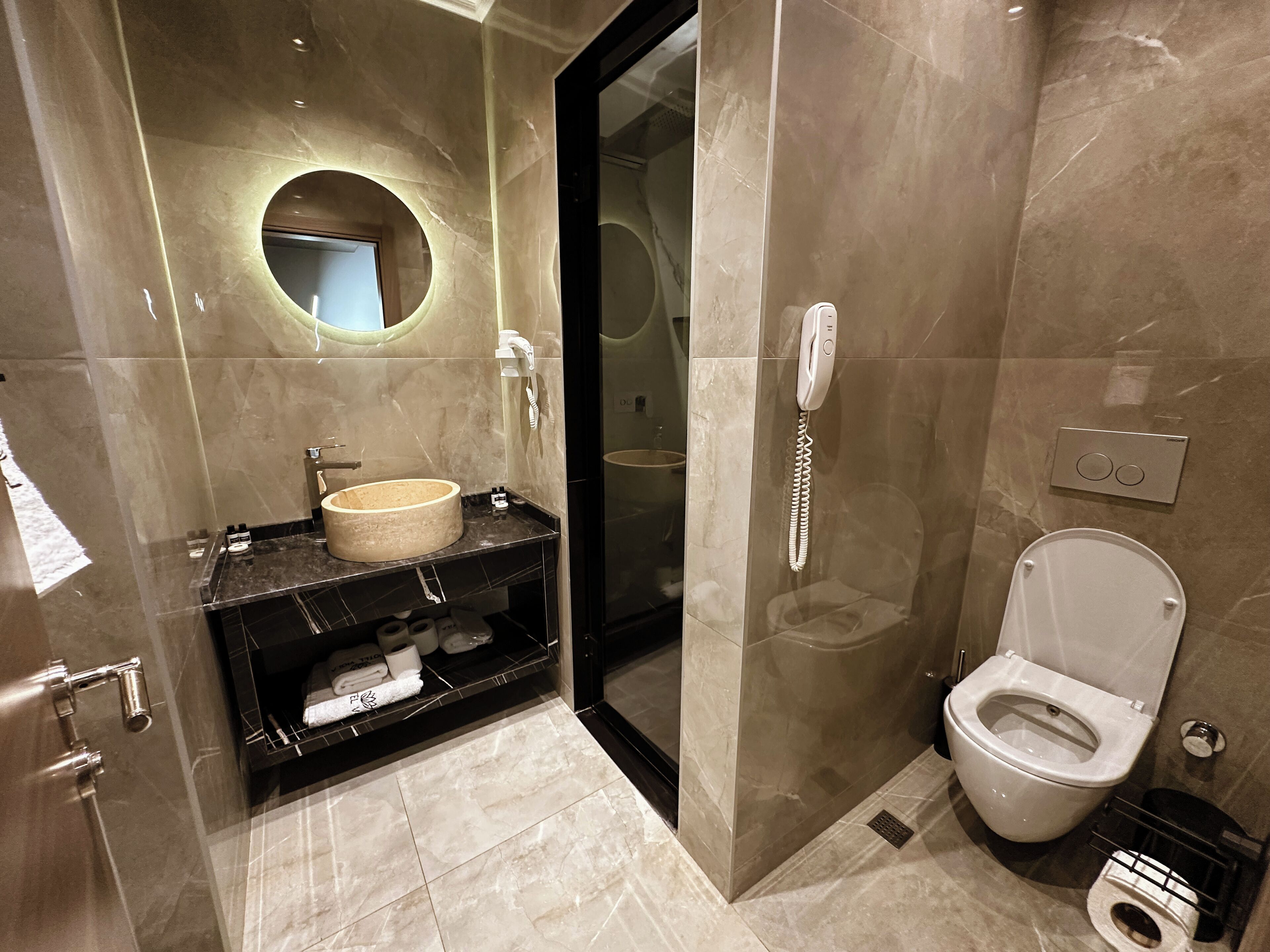 Een moderne badkamer met een ronde spiegel, een beige wastafel, toilet en een douchecabine met glazen wanden.