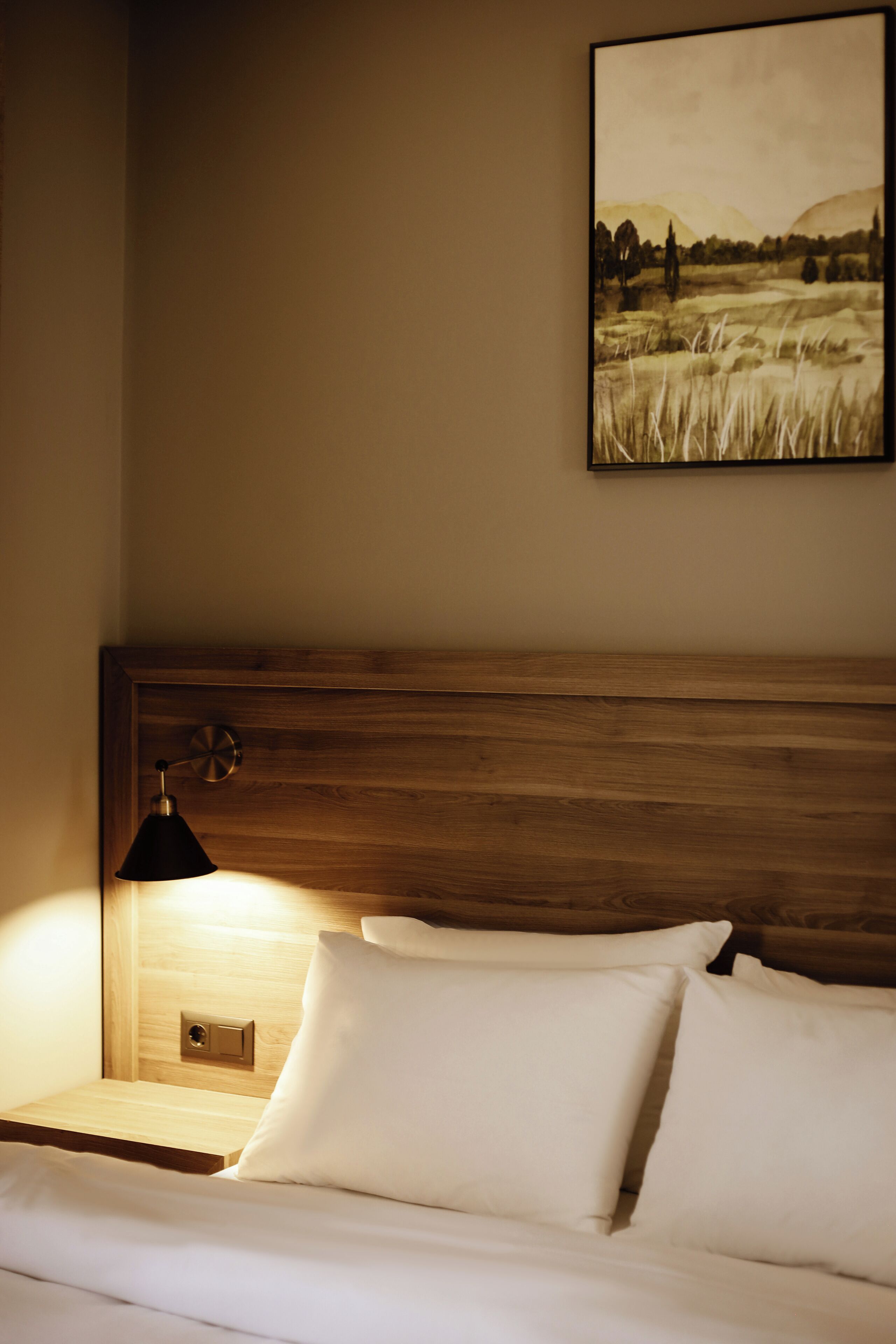 A neatly made bed with white linens, a wooden headboard, and a wall-mounted lamp.