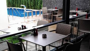 An outdoor dining area with tables and chairs, a pool in the background, and a stone wall.