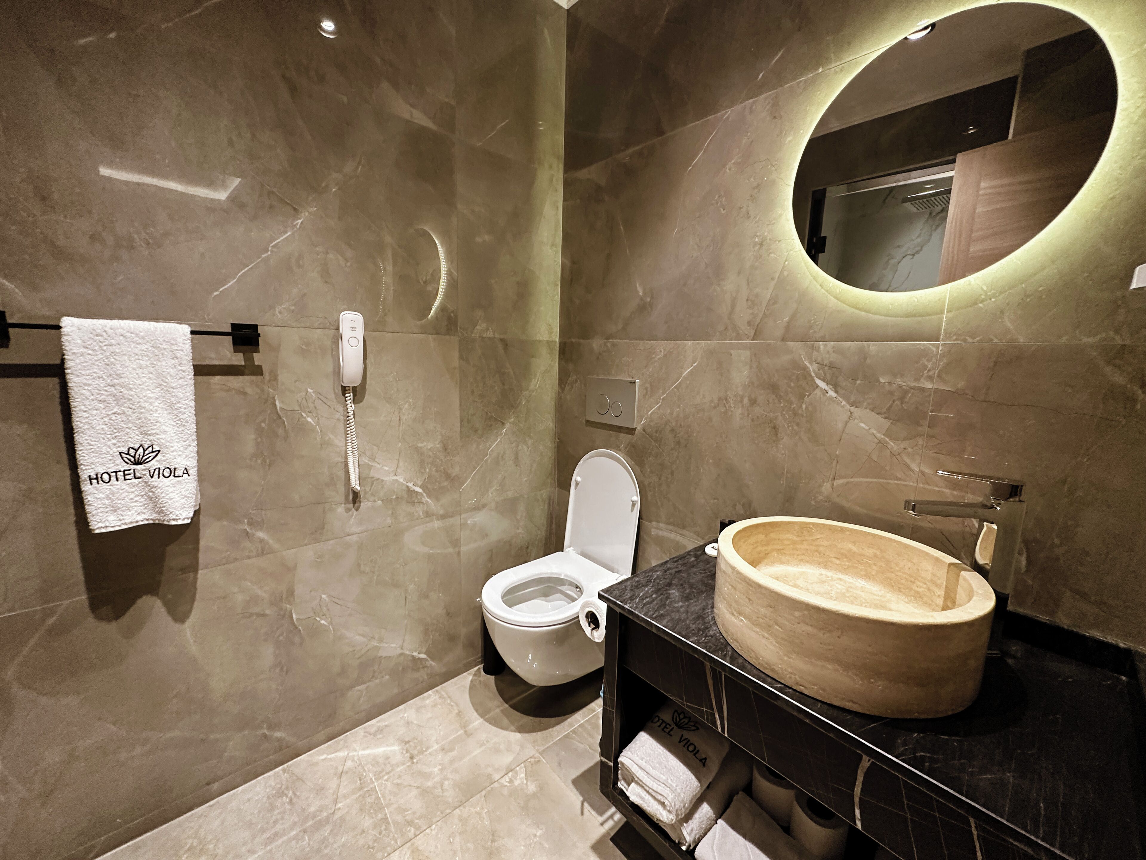 Un baño moderno con suelo de mármol, lavabo de madera, inodoro y una toalla colgada en un perchero.