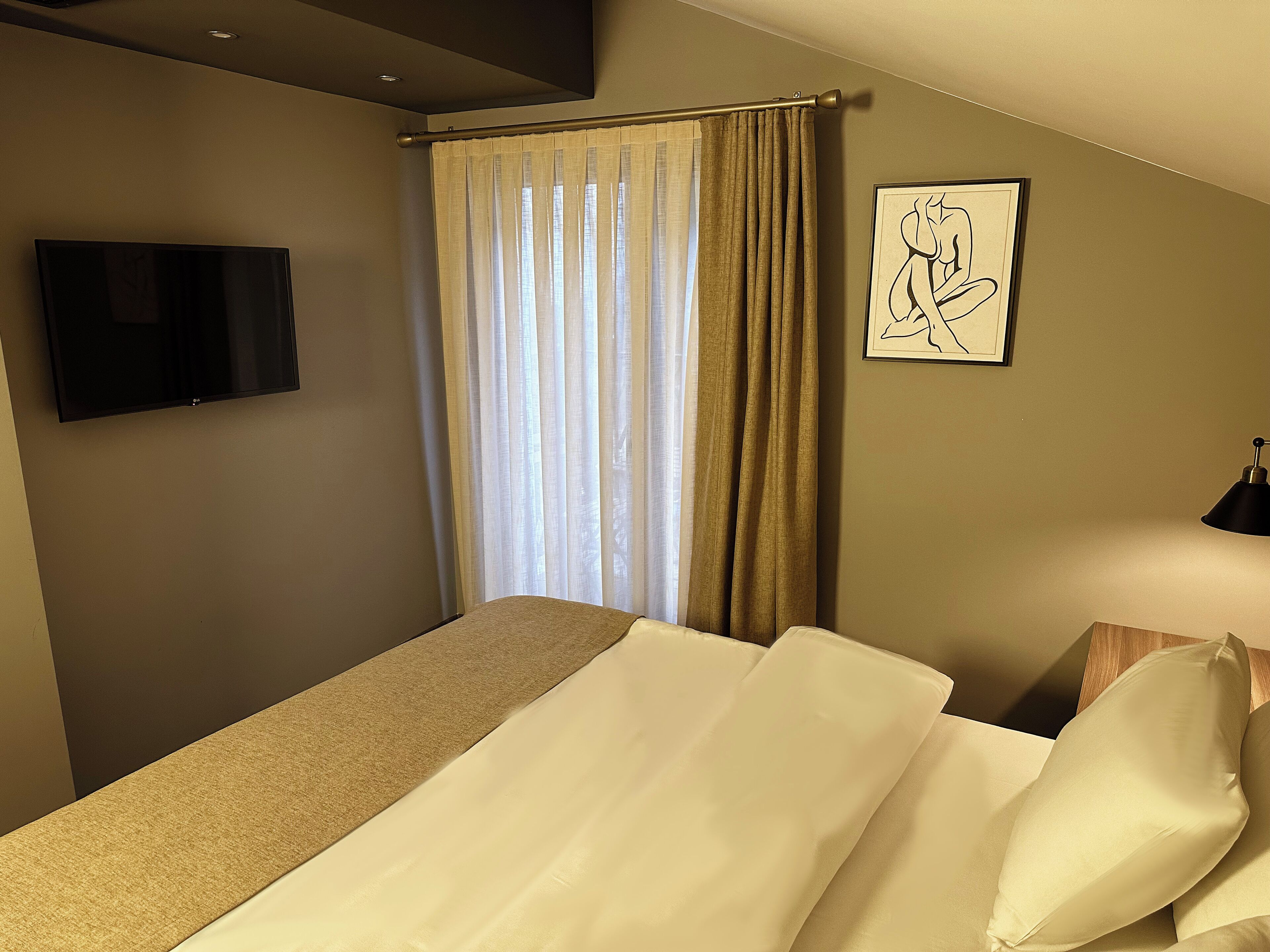 A hotel room with a bed, a TV, a window with curtains, and a framed picture on the wall.