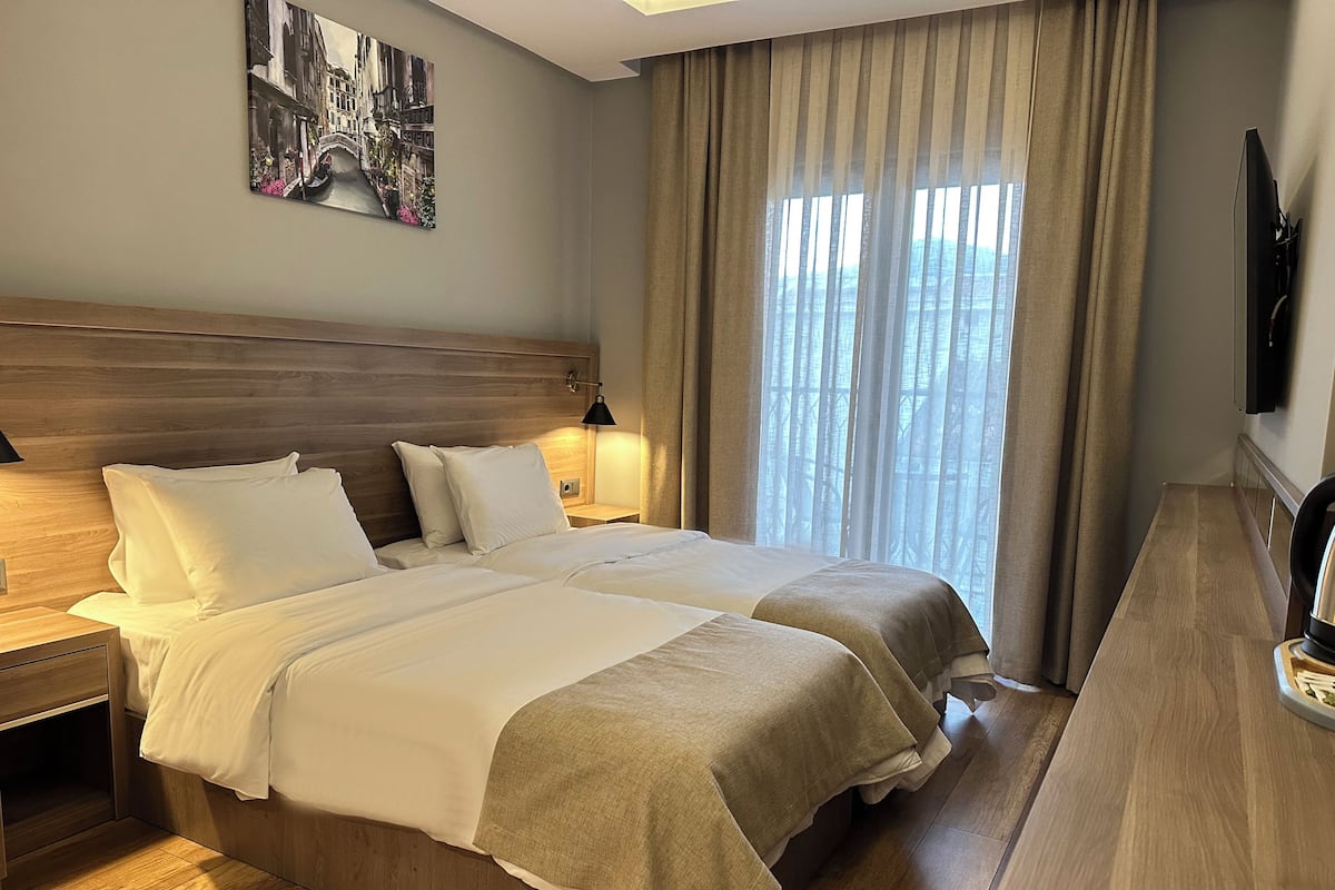 Une chambre d’hôtel avec deux lits, une tête de lit en bois, un tableau au mur, une télévision et une fenêtre avec des rideaux.