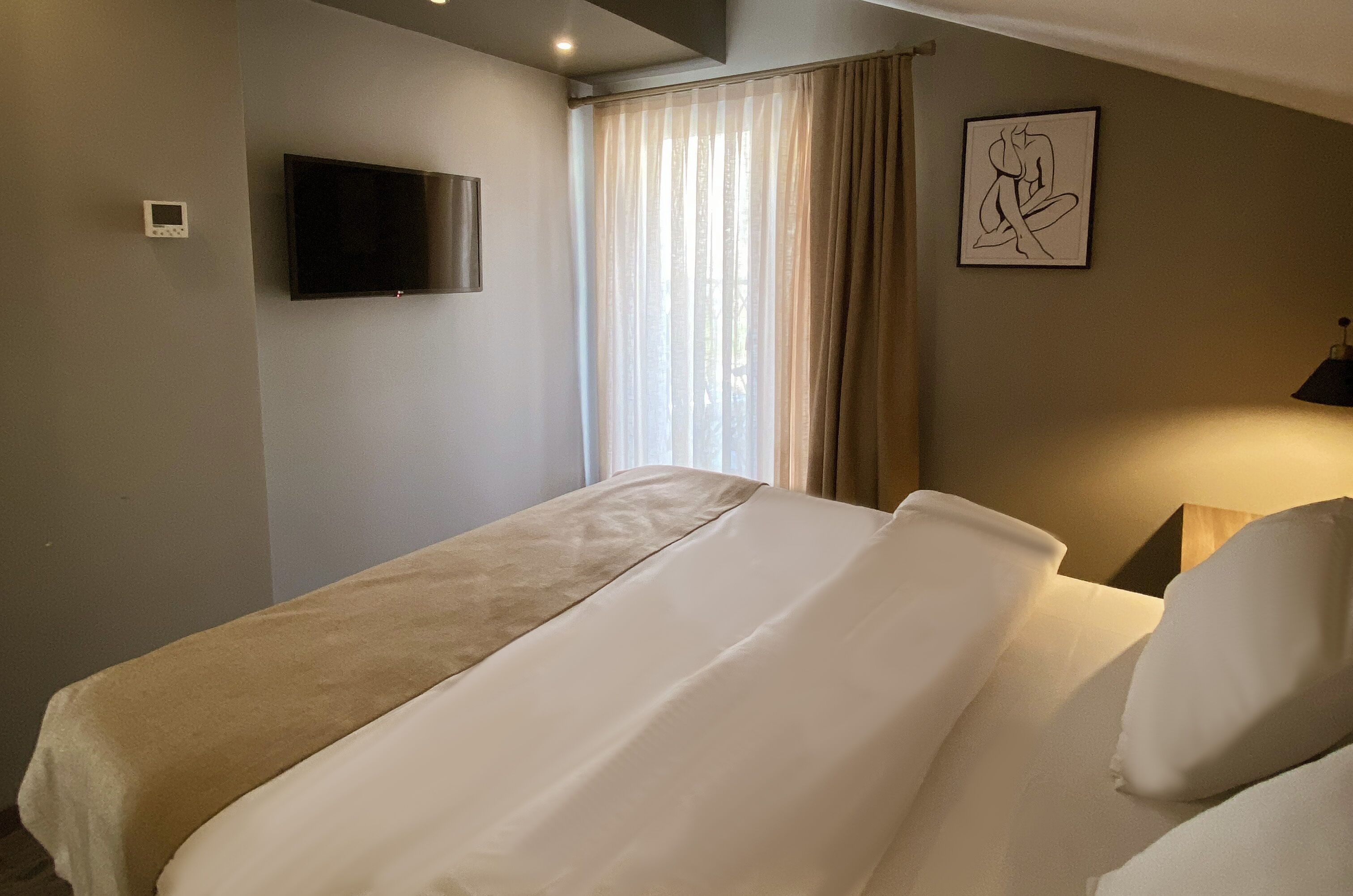 Habitación de hotel con cama, televisor de pantalla plana en la pared, ventana con cortinas translúcidas y un cuadro enmarcado.