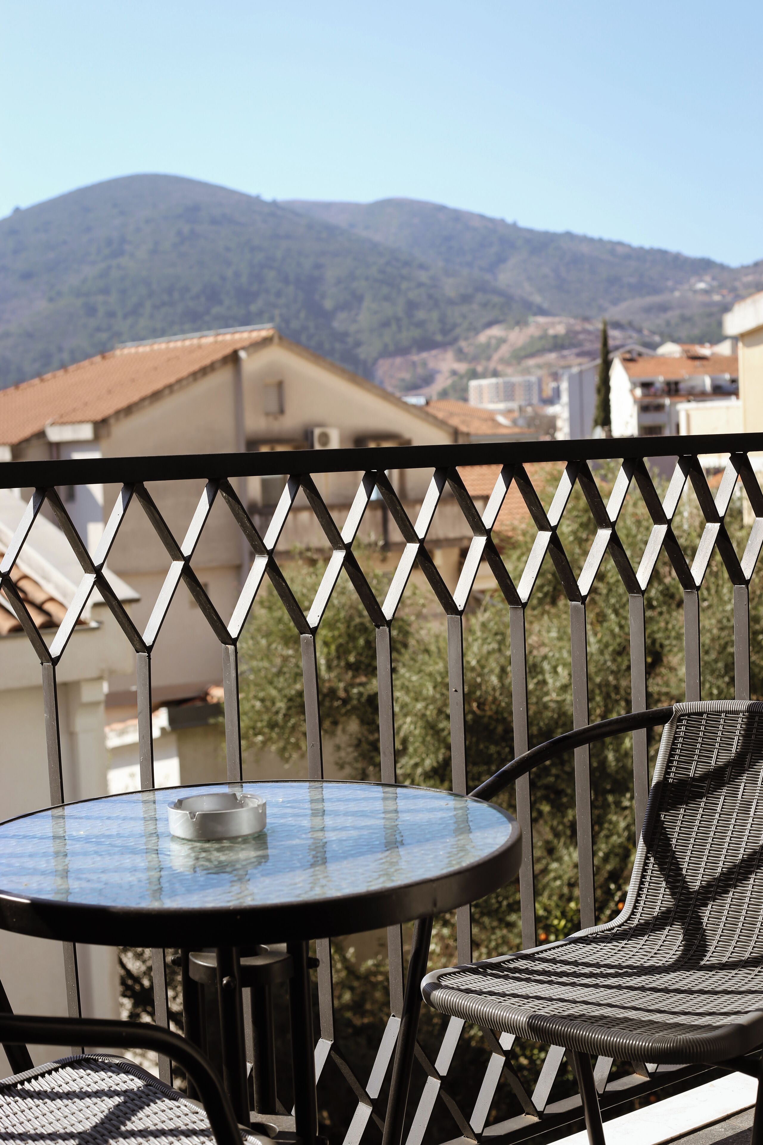 A balcony with a round glass table and a wicker chair, overlooking a mountainous landscape.