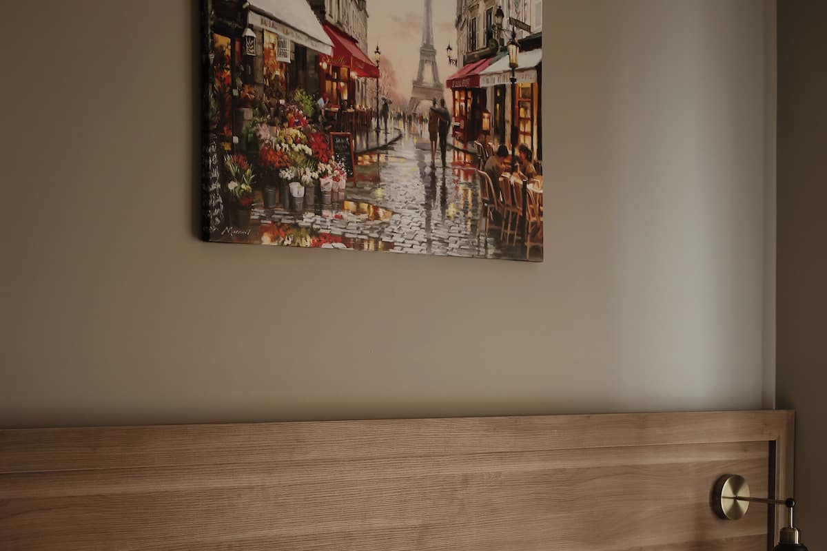 Une chambre d’hôtel avec une tête de lit en bois, du linge de lit blanc, un tableau représentant une scène de rue parisienne et une lampe de chevet.