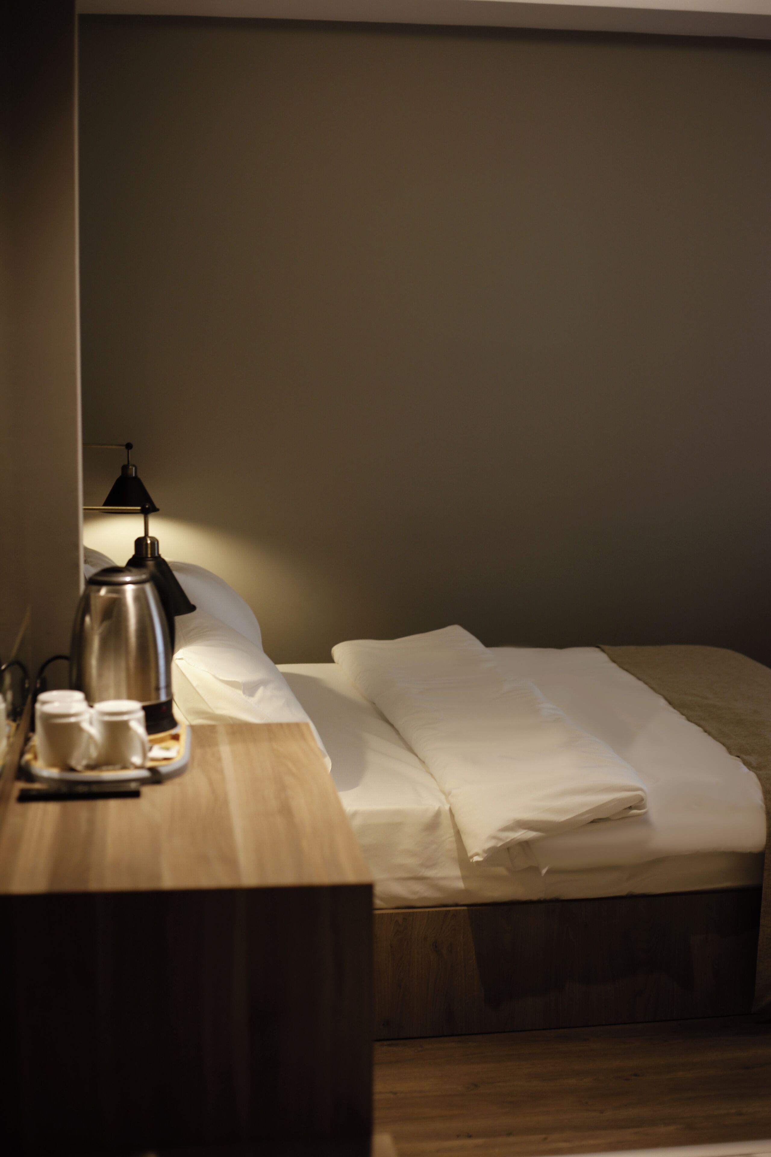 A neatly made bed with white linens, a bedside table with a coffee pot, cups, and a lamp.