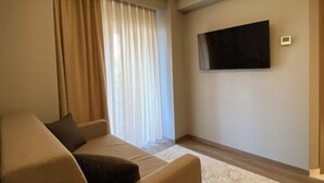 A modern living room with a sofa, a flat-screen TV mounted on the wall, and a large window with curtains. - Viola Hotel Budva (Budva)