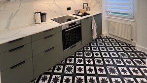 A modern kitchen with grey cabinets, a black and white patterned floor, and a window with blinds. - Appartement Familial 6 Personnes (Nancy)
