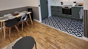 A modern kitchen with a built-in oven, microwave, and grey cabinets. There is a dining area with a table and chairs, and a patterned floor. - Appartement Familial 6 Personnes (Nancy)