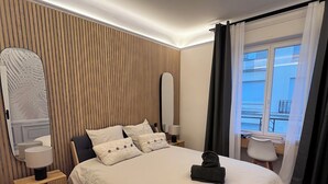 A modern bedroom with a large bed, wooden panel wall, bedside tables, and a view of the outside. - Appartement Familial 6 Personnes (Nancy)