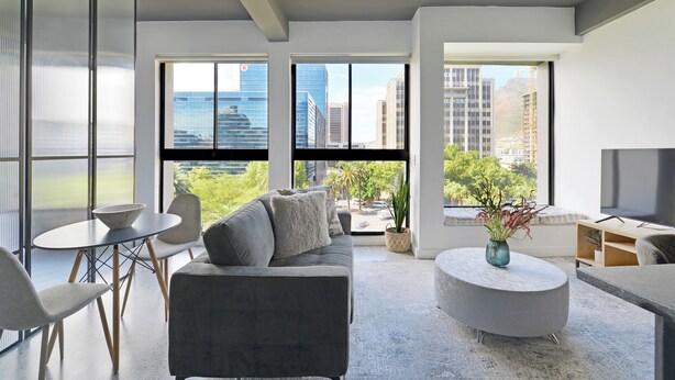 A modern living room with a grey sofa, round coffee table, and a dining table with chairs. Large windows offer views of the cityscape and greenery. - Modern City Escape with Rooftop Pool Views (Cape Town)