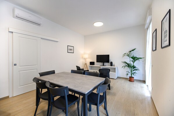 En moderne stue med spisebord, stole, et fladskærms-tv, en lampe og en potteplante.