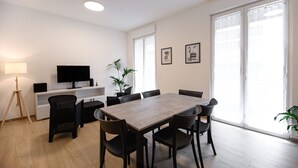 A modern living room with a wooden dining table, black chairs, a flat-screen TV on a white stand, a floor lamp, and two potted plants. - Ca dei Gianchi 3 Apartment with 2 rooms and private parking with video surveillance (La Spezia)