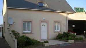 A two-story house with a beige exterior, a white door, and two windows with shutters. There is a satellite dish on the left side and a garage on the right.