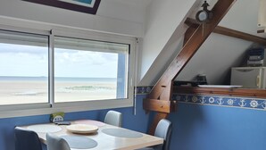 A dining area with a table and chairs, a view of the beach, and a clock on the wall.
