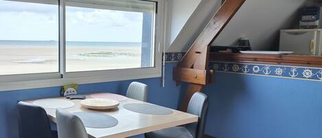 A dining area with a table and chairs, a view of the beach, and a clock on the wall.