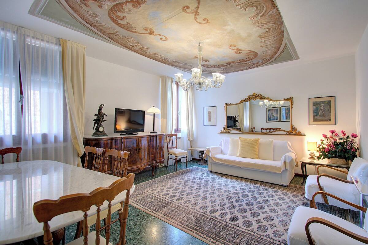 A spacious living room with a marble table, wooden chairs, a white sofa, a chandelier, and a large mirror.