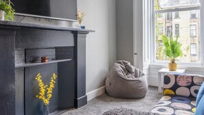 A modern living room with a fireplace, a bean bag, a sofa with a colorful pattern, and a view of buildings through a large window. - West End Cosy Pop of Colour - Pass the Keys (Glasgow)