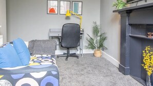A modern office space with a grey sofa, a black office chair, a desk with a computer, a yellow desk lamp, framed artwork on the wall, a small potted plant, and a fireplace. - West End Cosy Pop of Colour - Pass the Keys (Glasgow)