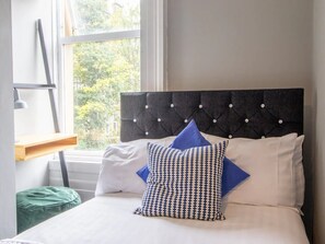 Una cama bien hecha con cabecera capitonada, una mesita de noche con lámpara y vistas a la vegetación por la ventana.