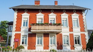 En rød murbygning med hvide detaljer, mange vinduer og en balkon med planter.