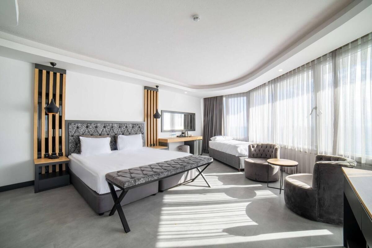 Una habitación de hotel moderna con una cama grande, un escritorio, una silla y un sofá. Hay una ventana con cortinas y vistas al exterior.