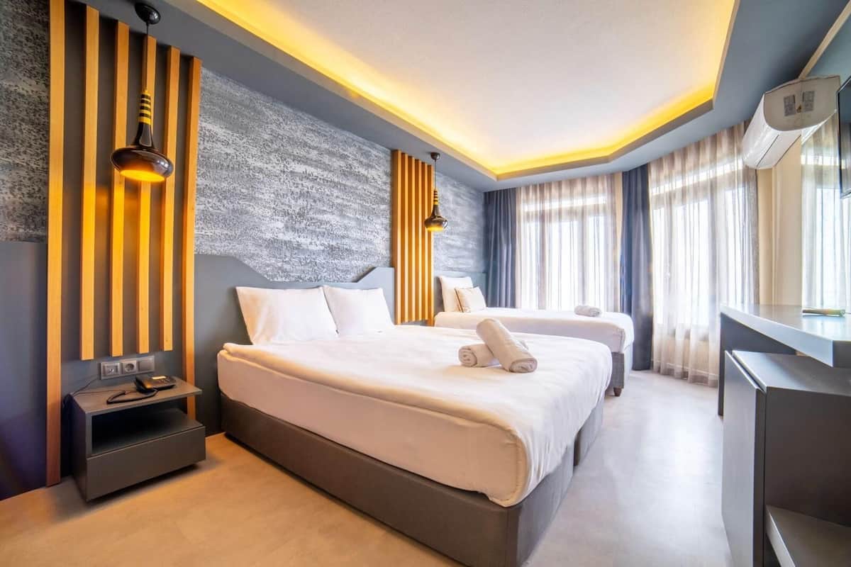 Una habitación de hotel moderna con una cama grande, mesitas de noche, un escritorio y un ventanal con cortinas.