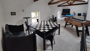 Un grand espace de vie avec une table à carreaux, un canapé et une table de baby-foot. On aperçoit une télévision et une table de billard au fond.