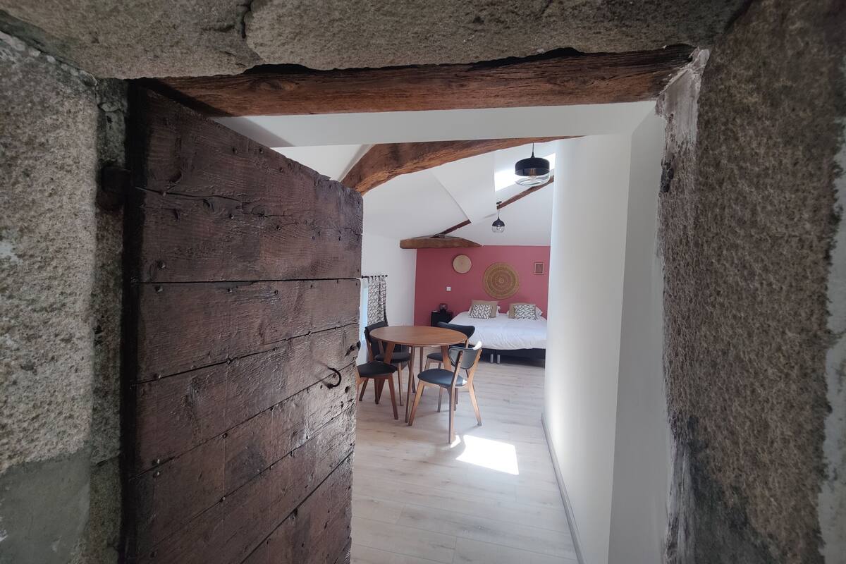 Quarto rústico com vigas de madeira aparentes, uma mesa redonda de madeira com cadeiras, uma cama com roupa de cama estampada e um painel com um objeto circular trançado.