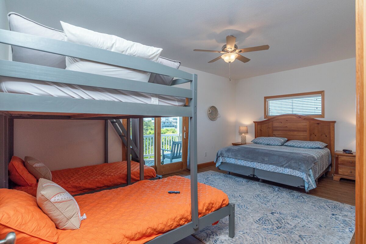 A room with bunk beds, a single bed, a ceiling fan, a window with blinds, a wooden nightstand, and a patterned rug.