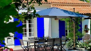 Un área de comedor al aire libre con una mesa y sillas, una sombrilla azul y un edificio de paredes azules y blancas.