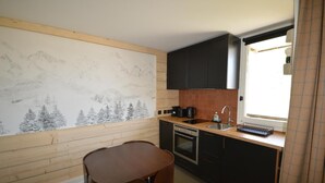 A compact kitchen with a wooden wall mural, a sink, a stove, and a dining area with a table and chairs. - 30m from ski lifts, 2nd floor, view ski slopes, terrace, tv, ski locker, 19m², Montalbert (La Plagne)