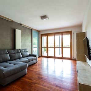 Amplia sala de estar con sofá seccional gris, piso de madera, grandes puertas correderas de vidrio y televisor de pantalla plana montado en la pared.