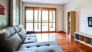 A modern living room with a grey sectional sofa, a wooden floor, a large sliding glass door leading to an outdoor area, and a built-in TV shelf. - Cave 47 Zona Ospedale Giovanni Xxiii (Bergamo)
