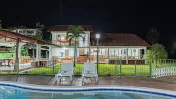 A swimming pool area with lounge chairs, a covered seating area, and a building with a tiled roof.