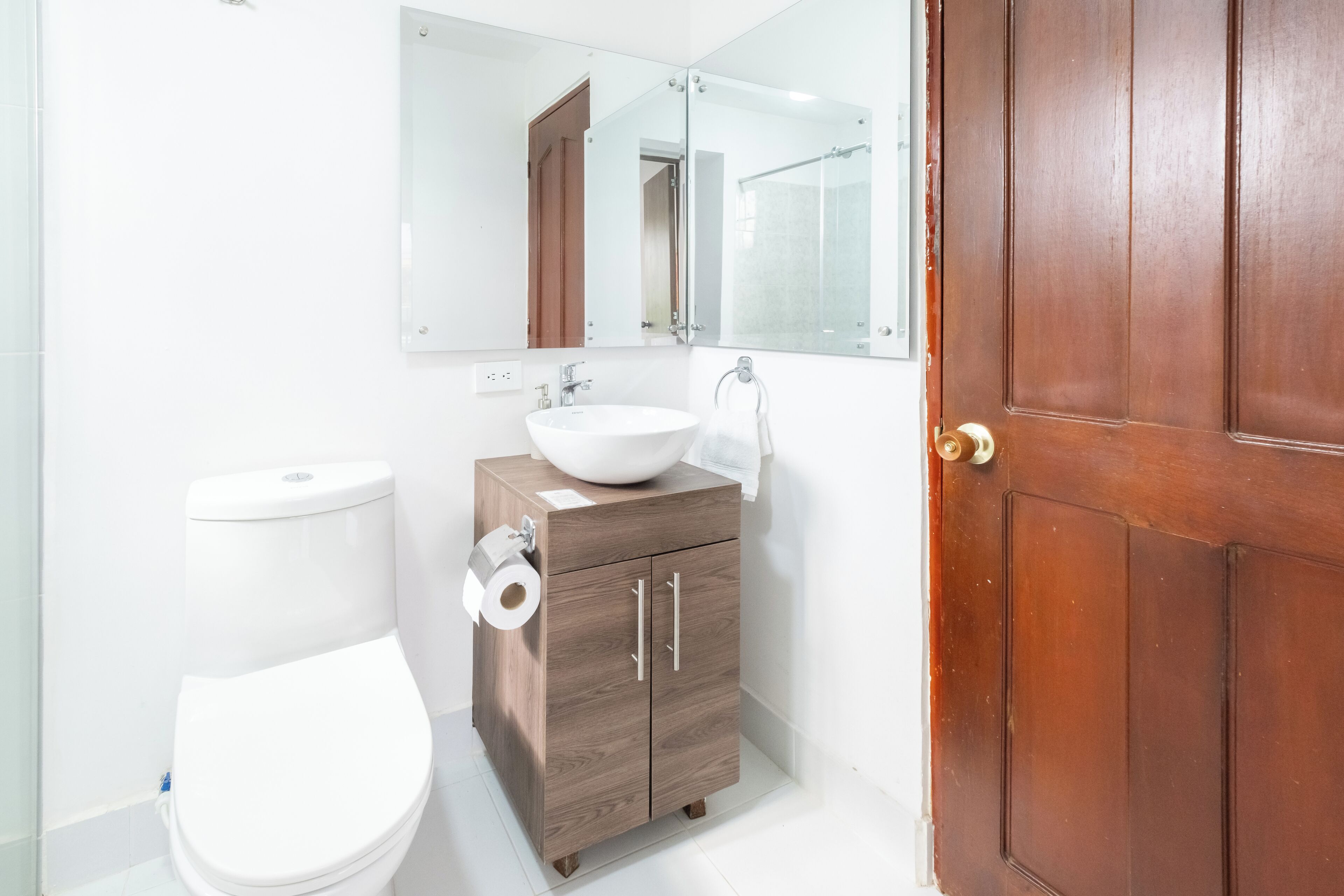A bathroom with a white toilet, a wooden cabinet with a sink, and a large mirror above the mirror cabinet.
