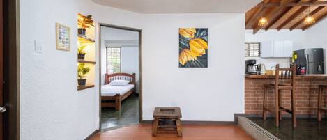 A compact living space with a kitchen area, a small stool, a bed, and a door leading to another room.