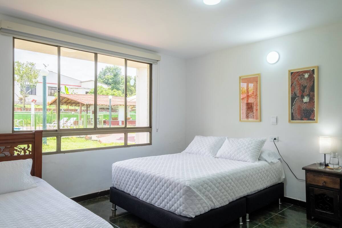 Quarto com uma grande janela, uma cama, uma mesa de cabeceira e duas molduras na parede.