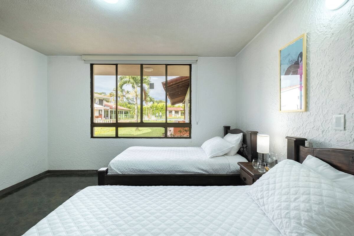 Quarto de hotel com duas camas, janela com vista para palmeiras e casas, mesa de cabeceira com abajur e quadro na parede.