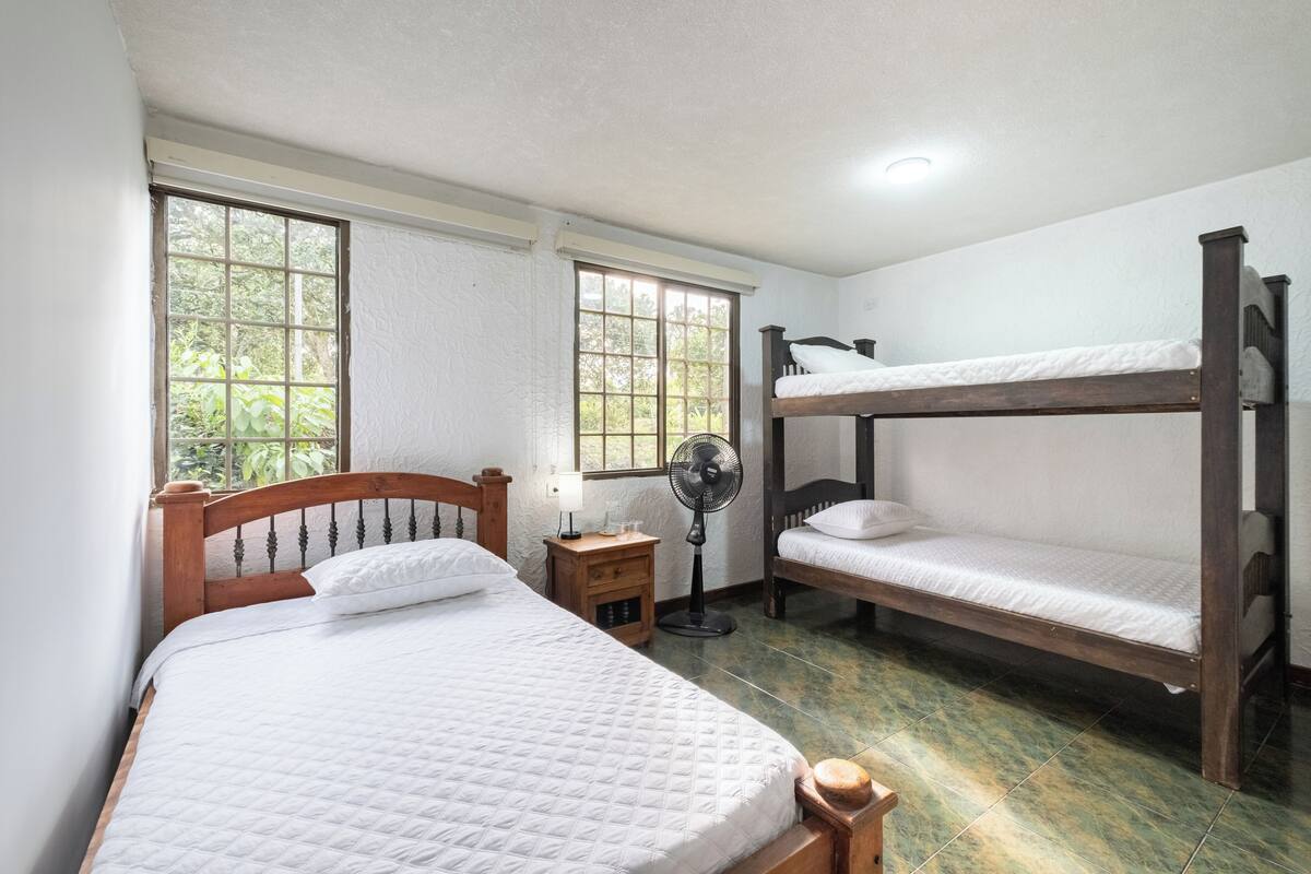 Um quarto de dormitório com uma cama de solteiro, uma cama em beliche, uma mesinha de cabeceira com abajur e um ventilador.