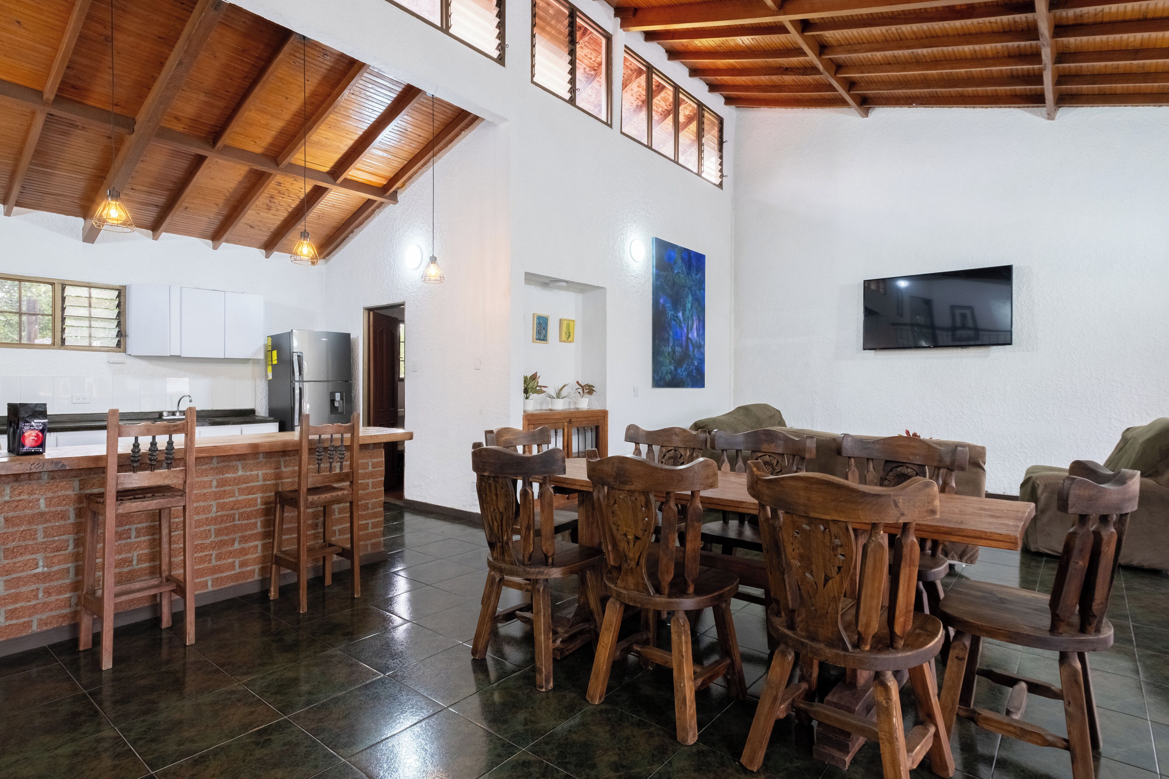 A spacious kitchen with a high ceiling and exposed beams, a dining area with wooden chairs and a table, and a flat-screen TV mounted on the wall.