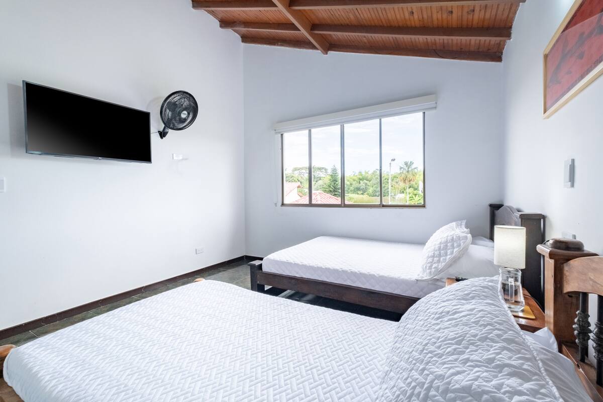 Quarto com duas camas, televisão fixada na parede, janela com vista e ventoinha no teto.