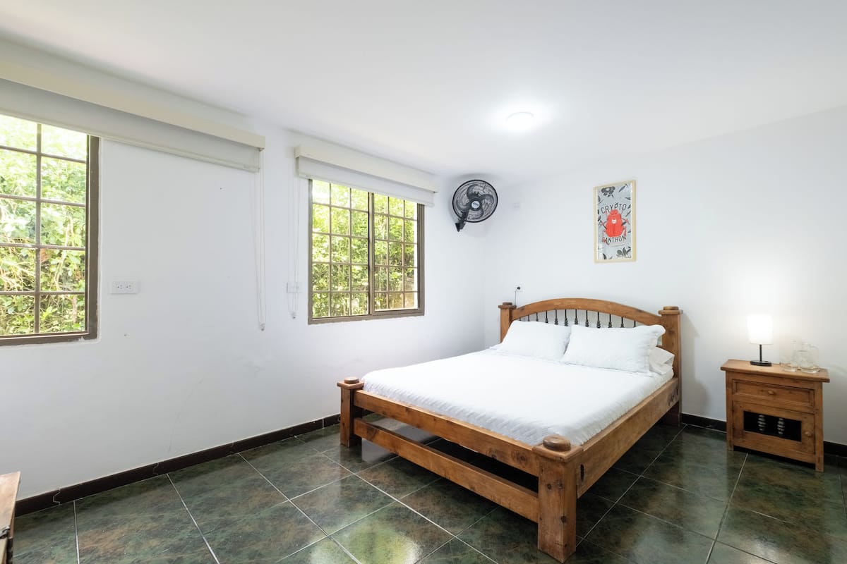 Quarto com cama de madeira, mesa de cabeceira, ventilador de parede e duas janelas.