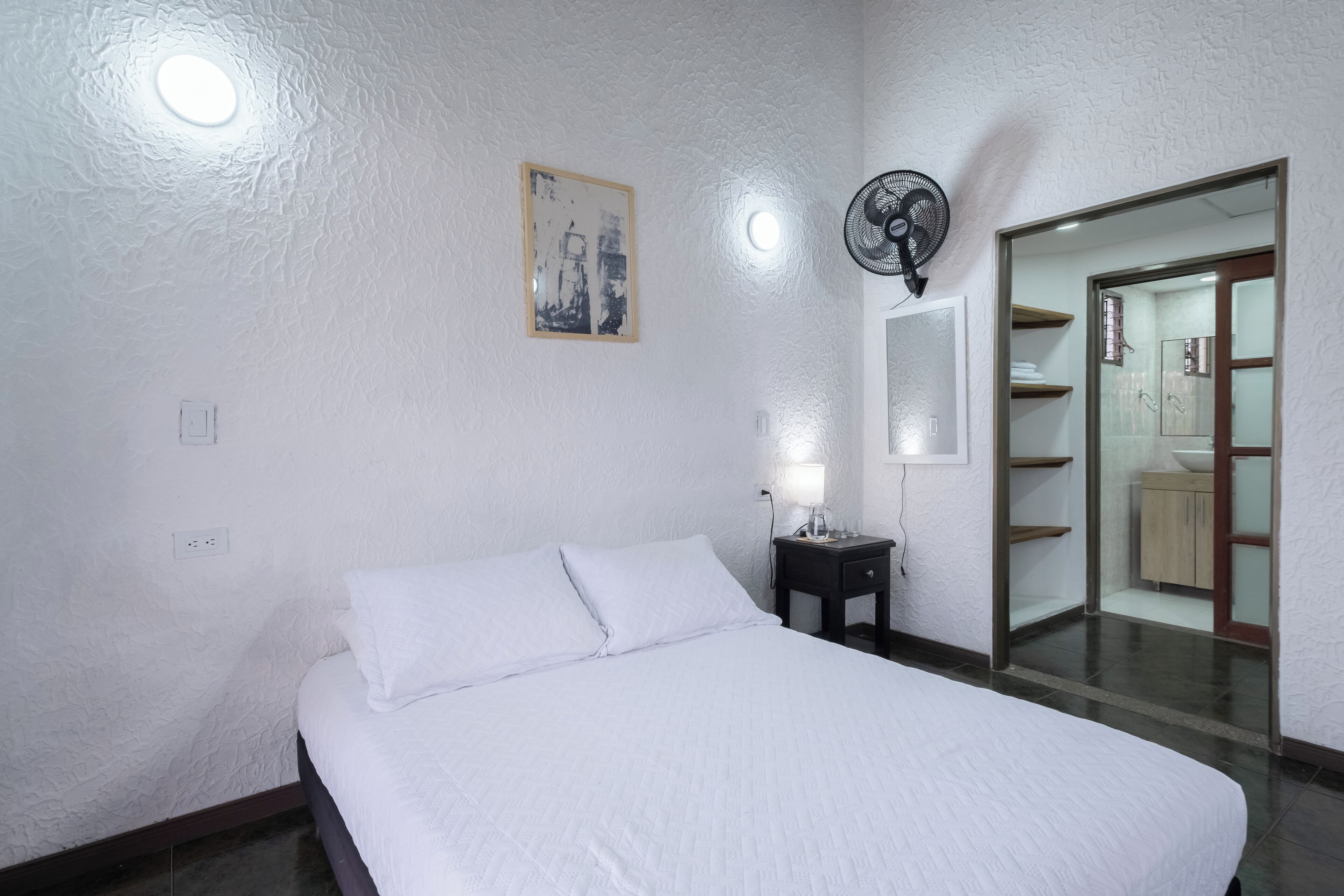 A neatly made bed in a room with a white textured wall, a small bedside table with a lamp, a ceiling fan, and a mirror reflecting a bathroom with shelves and a sink.