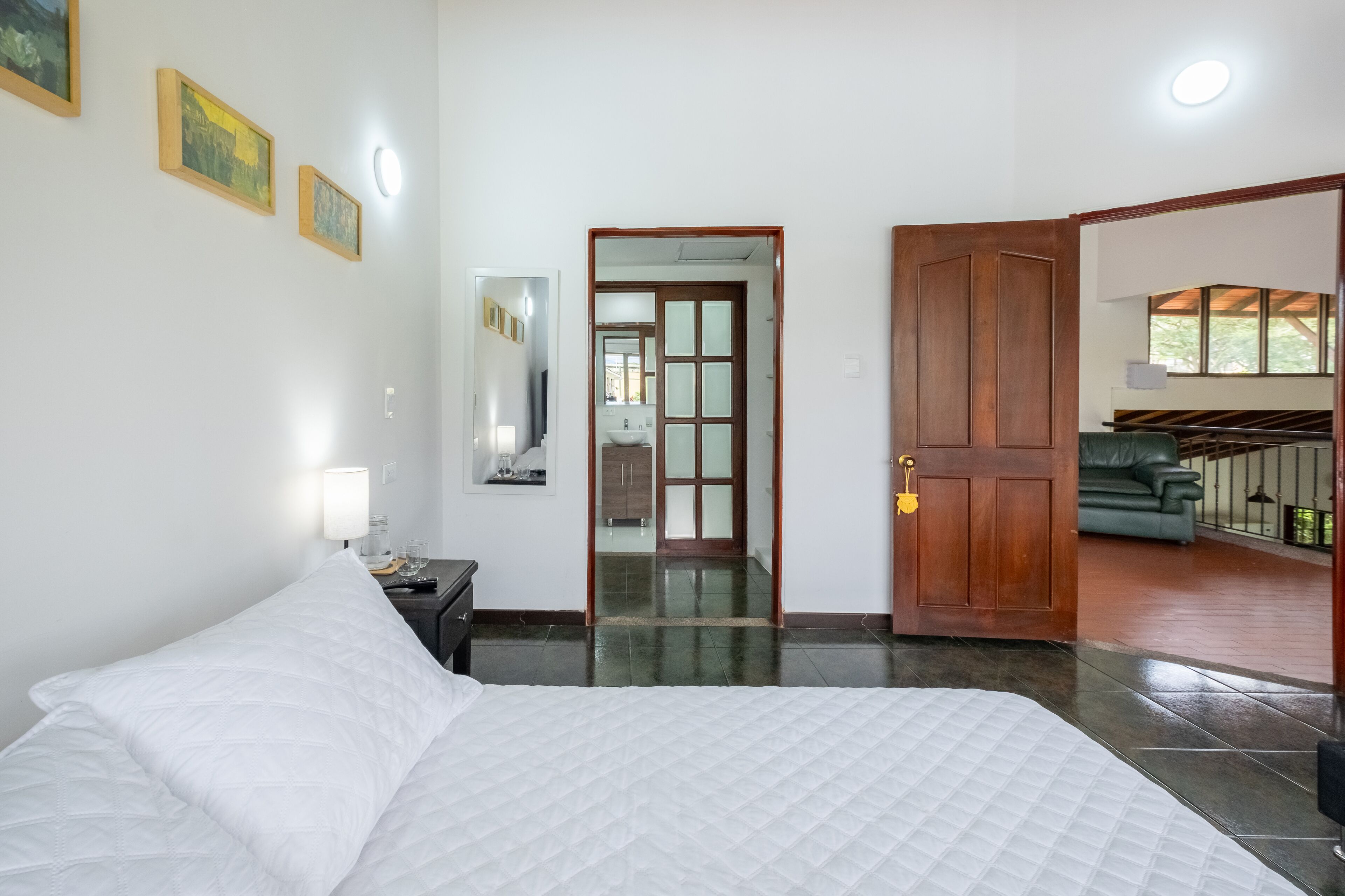 Quarto com cama, mesa de cabeceira, espelho e porta que dá para uma sala de estar.