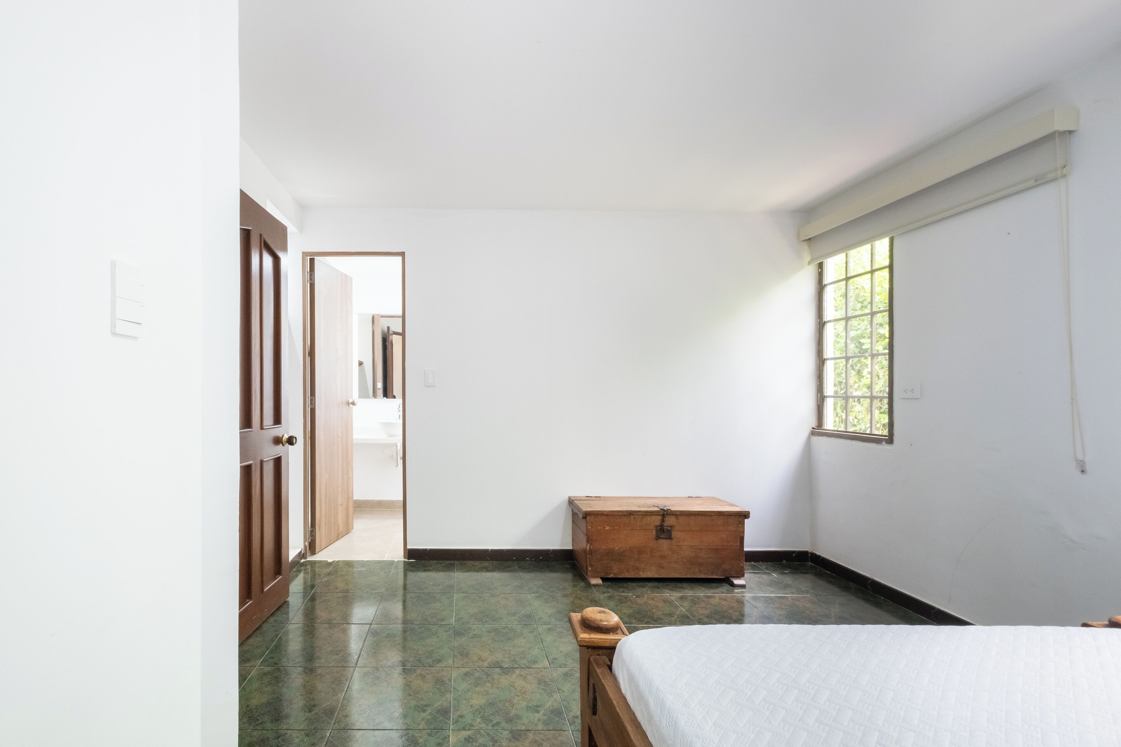 Ein Schlafzimmer mit Bett, einem hölzernen Truhe, einem Fenster und einer Tür zu einem anderen Raum.
