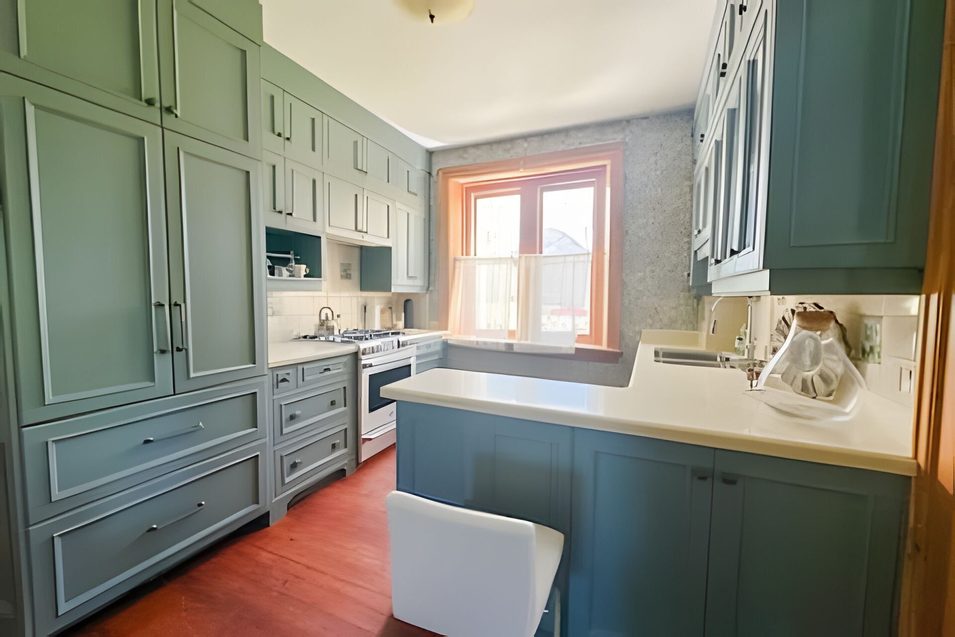 Una cocina con gabinetes verde claro, encimera blanca y una ventana con cortinas anaranjadas.
