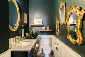 A bathroom with a white sink, a toilet, and two gold-framed mirrors. There are plants and a framed picture on the wall.